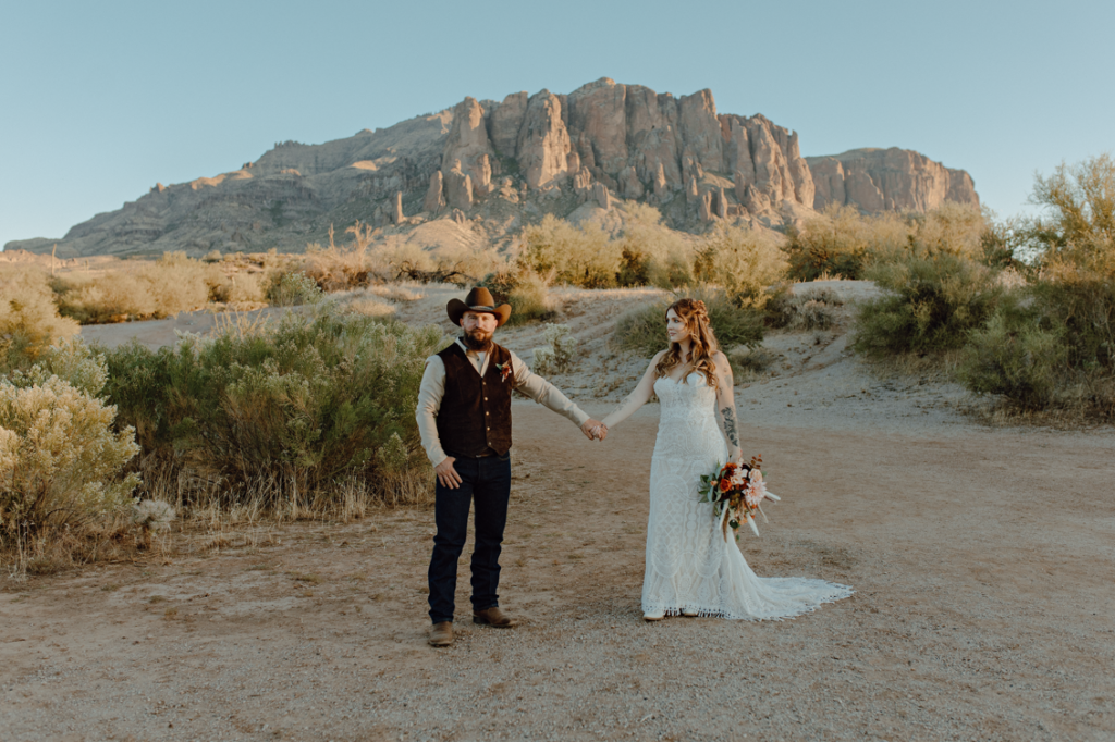 a couple holding hands in front of superstition mountains while taking wedding portraits. 