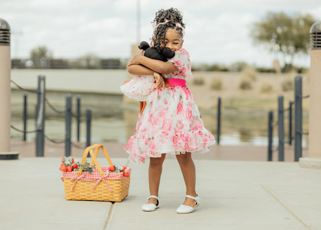 A little girl holding and hugging her doll in the park, standing next to a basket that has strawberries 