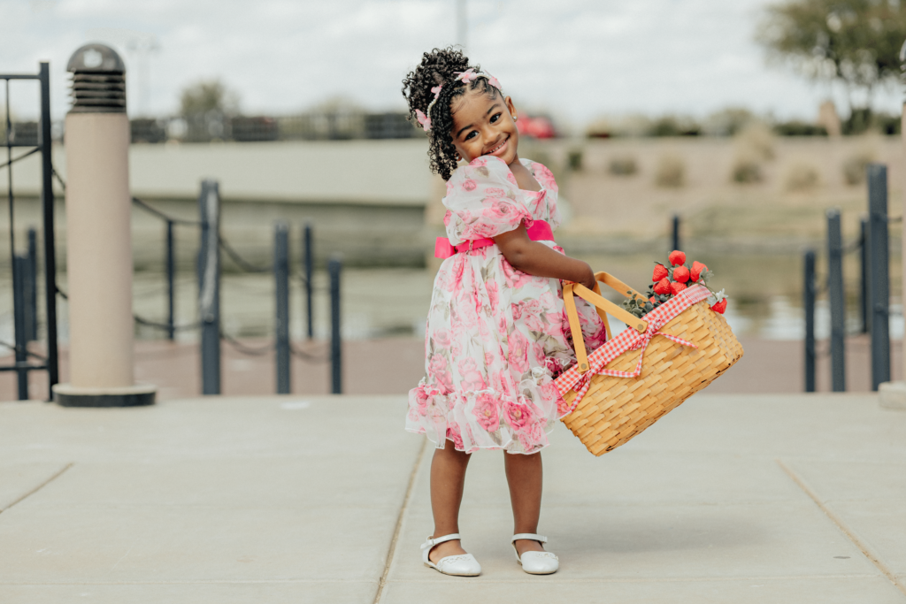 A little girl holding her basket that has strawberries and smiling. she's has on a colorful flower pink dress