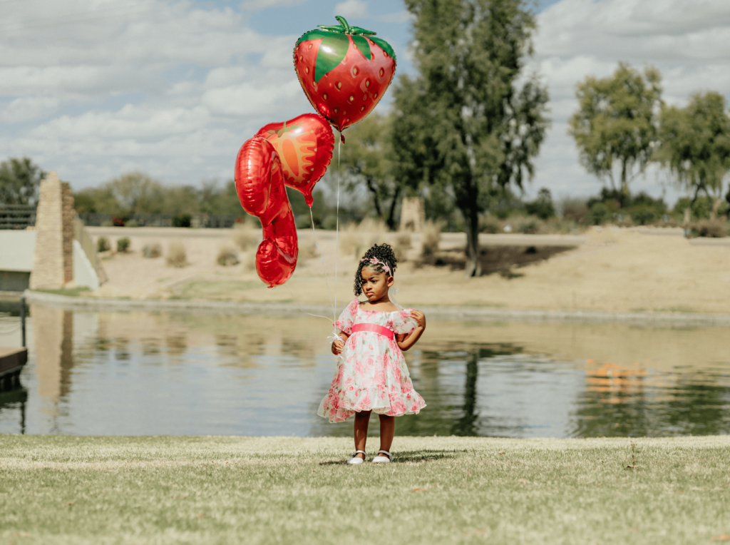 a little girl holding balloons in the park while there's a lake behind her