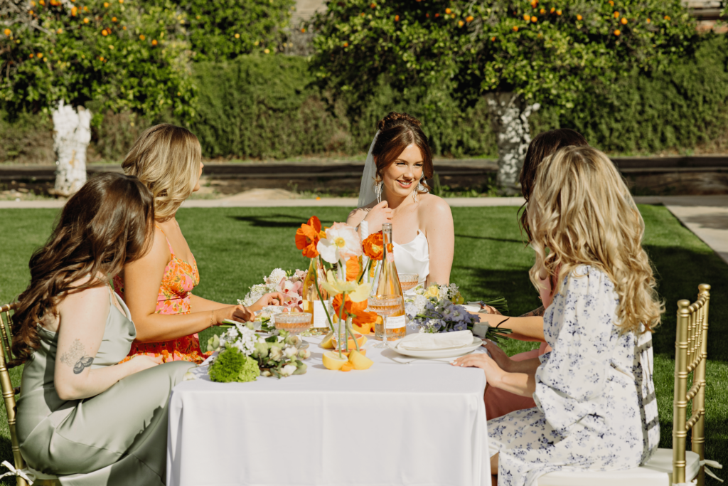Bride sitting on a table around her bridesmaid. they are wearing colorful spring dresses and the table is covered with floral centerpieces.