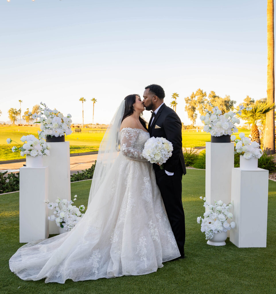 A bride and groom share a kiss outdoors, surrounded by white floral arrangements. The bride wears a flowing lace gown and veil, conveying romance and joy.